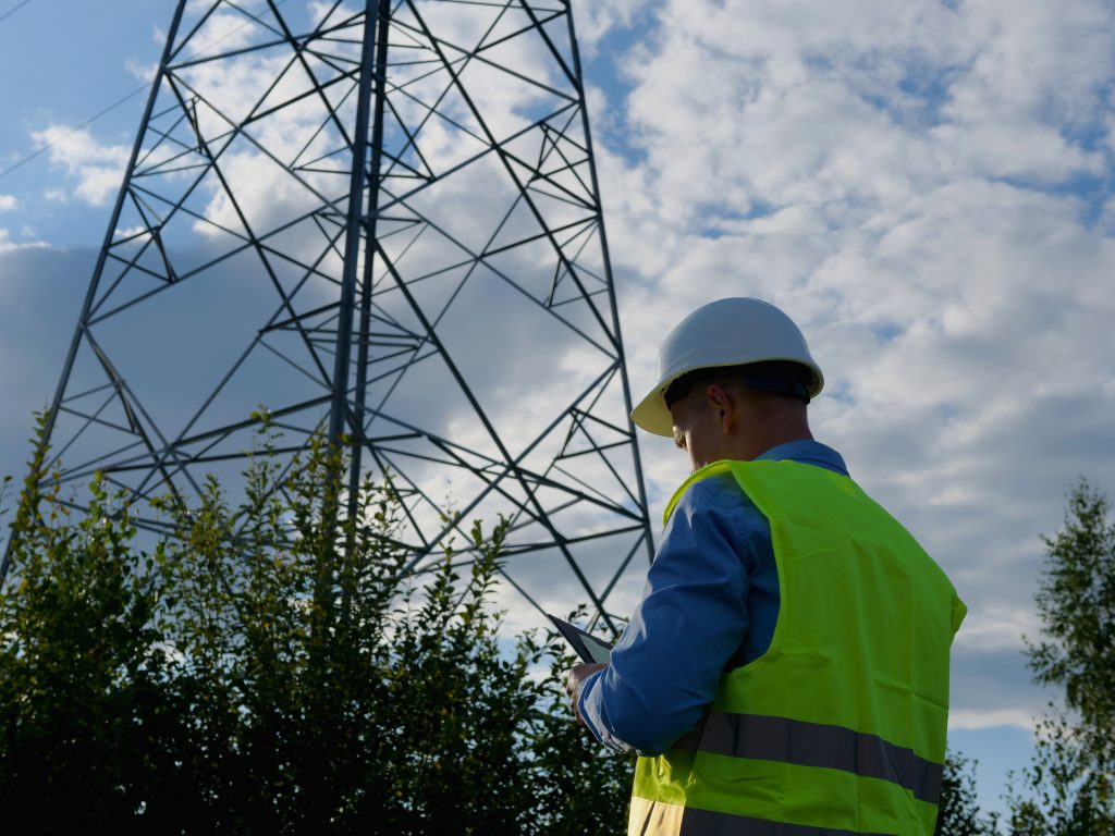Equipped with a white helmet, reflective vest, and tablet, an energy engineer examines an electric tower and wires. This energy engineer is committed to maintaining power distribution systems.